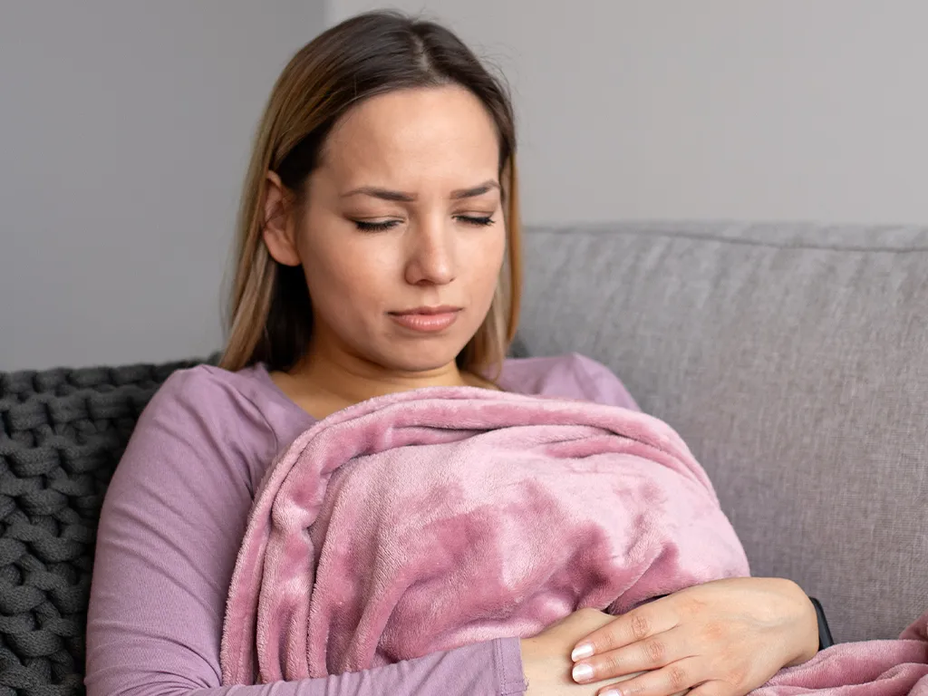 Woman sitting on a couch under a blanket, holding her abdomen in discomfort
